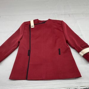Op de afbeelding: Bordeaux jasje met zwarte bies en een crèmekleurig accent op de schouder en mouw. De jas heeft een asymmetrisch ontwerp met een sluiting met één knoop en twee kleine zakken. De mouwen zijn lang en de jas lijkt van een gladde stof te zijn.