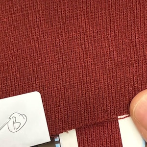 May include: Close-up of a section of rust-colored knit fabric. The fabric has a ribbed texture with vertical lines. A small white tag with a handwritten "B" is in the lower left corner. A finger is visible in the lower right corner.