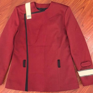 May include: A burgundy jacket with a black trim and a white stripe on the shoulder and sleeve. The jacket has a unique asymmetrical design with a black trim that runs down the front. The sleeve cuff has a white stripe.