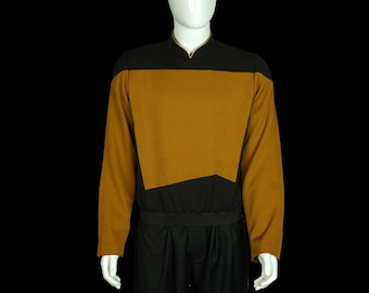 Puede incluir: Una camisa de manga larga con cuello negro y un panel de color dorado en la parte delantera. La camisa tiene una banda negra alrededor de la cintura.