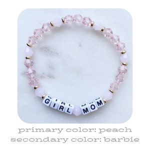 May include: A pink and gold beaded bracelet with the words "Girl Mom" spelled out in white letter beads.