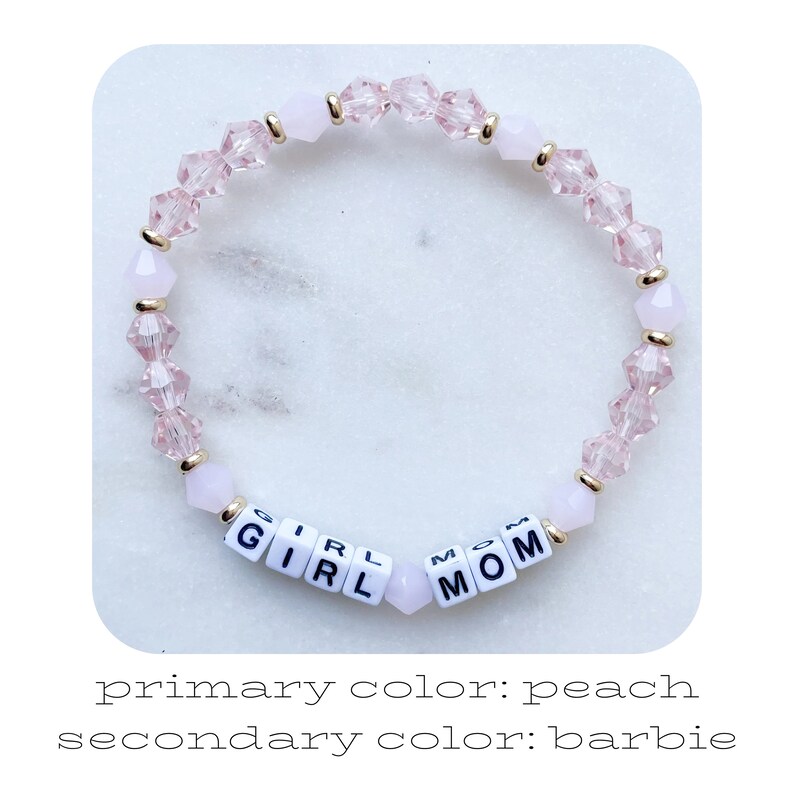 May include: A pink and gold beaded bracelet with the words "Girl Mom" spelled out in white letter beads.