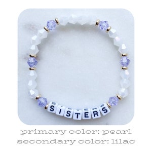 May include: A white beaded bracelet with lilac crystal accents and the word "SISTERS" spelled out in white letter beads.