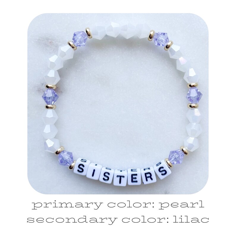 May include: A white beaded bracelet with lilac crystal accents and the word "SISTERS" spelled out in white letter beads.