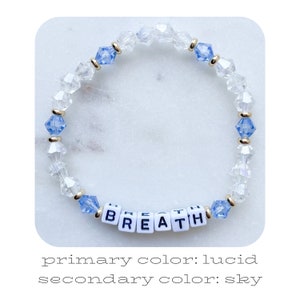 May include: A beaded bracelet with clear and sky blue crystals, spelling out the word "BREATHE" in white letters.