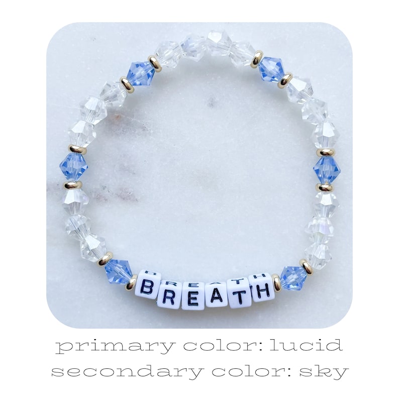 May include: A beaded bracelet with clear and sky blue crystals, spelling out the word "BREATHE" in white letters.