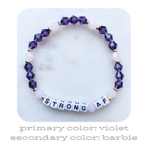 May include: A purple and pink beaded bracelet with gold accents. The bracelet has white letter beads that spell out "STRONG AF".