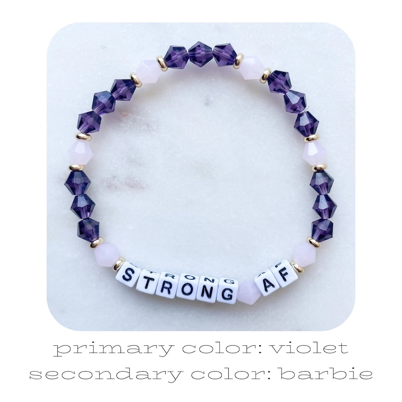 May include: A purple and pink beaded bracelet with gold accents. The bracelet has white letter beads that spell out "STRONG AF".