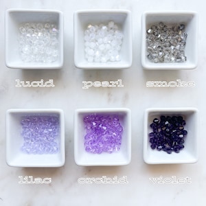 May include: Six small white bowls each containing a different colour of glass beads. The beads are labelled "lucid", "pearl", "smoke", "lilac", "orchid", and "violet".