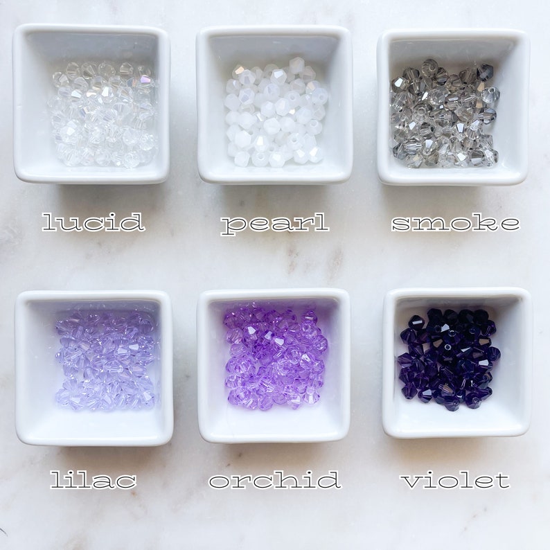 May include: Six small white bowls each containing a different colour of glass beads. The beads are labelled "lucid", "pearl", "smoke", "lilac", "orchid", and "violet".