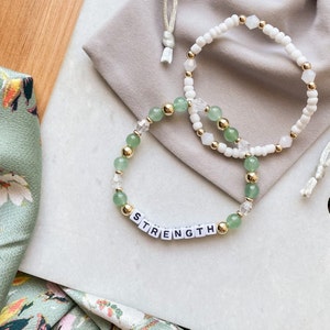May include: Two beaded bracelets. One bracelet features the word "STRENGTH" spelled out in white letter beads. The other bracelet is white. Both bracelets have gold-colored beads and green and clear beads.