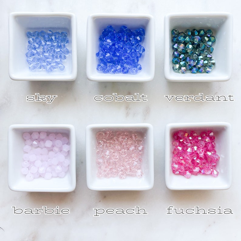 May include: Six small white bowls each containing a different colour of glass beads. The colours are sky blue, cobalt blue, green, light pink, peach, and fuchsia.