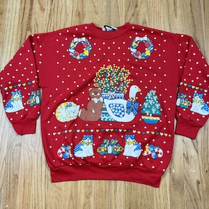 Puede incluir: Sudadera roja con temática navideña con lunares blancos. La sudadera presenta varios gráficos temáticos navideños, incluyendo un ganso, un oso de peluche, un árbol de Navidad, gatos y cajas de regalo. Las mangas tienen diseños de gatos y cajas de regalo.