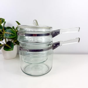 May include: A clear glass double boiler with a lid and two clear handles. The glass pot has a metal band around the middle and a clear glass lid with a small handle. The double boiler is sitting on a white surface.