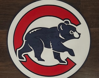 May include: A circular wooden wall sign featuring a large red letter C with a dark blue bear silhouette centered inside. The design is set against a white background with a dark blue outer border, creating a layered, multi-colored sports logo.