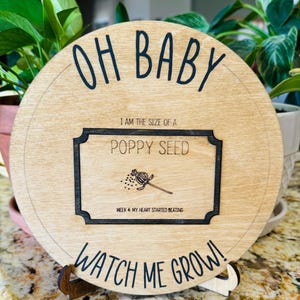 May include: A round wooden sign with the text "OH BABY" at the top and "WATCH ME GROW!" at the bottom. In the centre is a black and white image of a poppy seed with the text "I AM THE SIZE OF A POPPY SEED" and "WEEK 4: MY HEART STARTED BEATING".