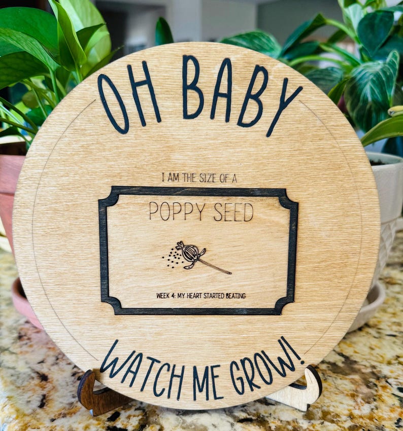 May include: A round wooden sign with the text "OH BABY" at the top and "WATCH ME GROW!" at the bottom. In the centre is a black and white image of a poppy seed with the text "I AM THE SIZE OF A POPPY SEED" and "WEEK 4: MY HEART STARTED BEATING".