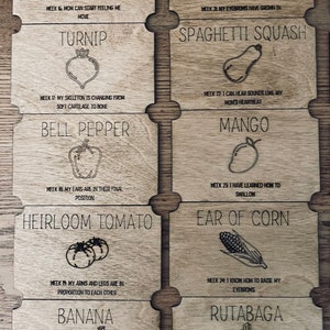 May include: A set of 10 wooden cards with illustrations of different fruits and vegetables. Each card has a title and a description of the stage of development of a baby during pregnancy. For example, the avocado card says "Week 16: Mum can start feeling me move".
