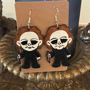 May include: A pair of brown and black Halloween-themed earrings featuring a cartoon character with a white mask and a knife. The earrings are hanging from a brown card.