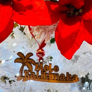 K&ouml;nnte beinhalten: Weihnachtsschmuck aus Holz mit den Worten "Mele Kalikimaka" und einem Palmenmotiv. Der Schmuck h&auml;ngt an einer rot-wei&szlig; gestreiften Perle und einem roten Band, vor einem wei&szlig;en Weihnachtsbaum mit roten Weihnachtssternen.