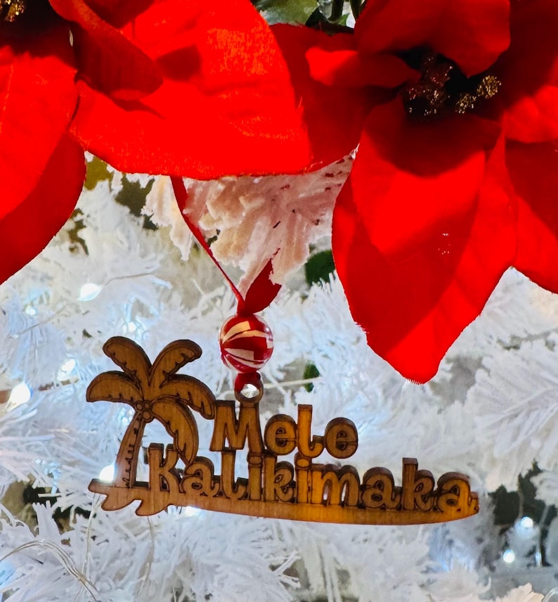 K&ouml;nnte beinhalten: Weihnachtsschmuck aus Holz mit den Worten "Mele Kalikimaka" und einem Palmenmotiv. Der Schmuck h&auml;ngt an einer rot-wei&szlig; gestreiften Perle und einem roten Band, vor einem wei&szlig;en Weihnachtsbaum mit roten Weihnachtssternen.