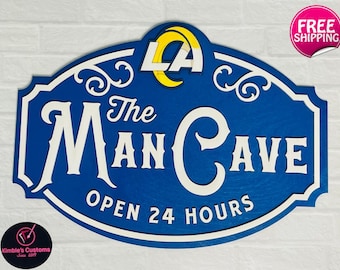 Könnte beinhalten: Ein blaues und weißes Holzschild mit dem Text "The Man Cave Open 24 Hours". Das Schild zeigt ein stilisiertes "C" und "M" im Text. Ein gelbes und blaues Logo mit den Buchstaben "LA" befindet sich oben in der Mitte des Schildes.