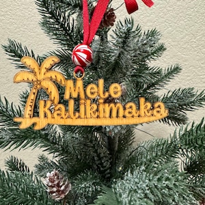 K&ouml;nnte beinhalten: Weihnachtsbaumschmuck aus Holz mit den Worten "Mele Kalikimaka" und einem Palmenmotiv. Der Schmuck h&auml;ngt an einem roten Band und ist mit einer rot-wei&szlig; gestreiften Kugel verziert. Der Schmuck wird auf einem Weihnachtsbaum pr&auml;sentiert.