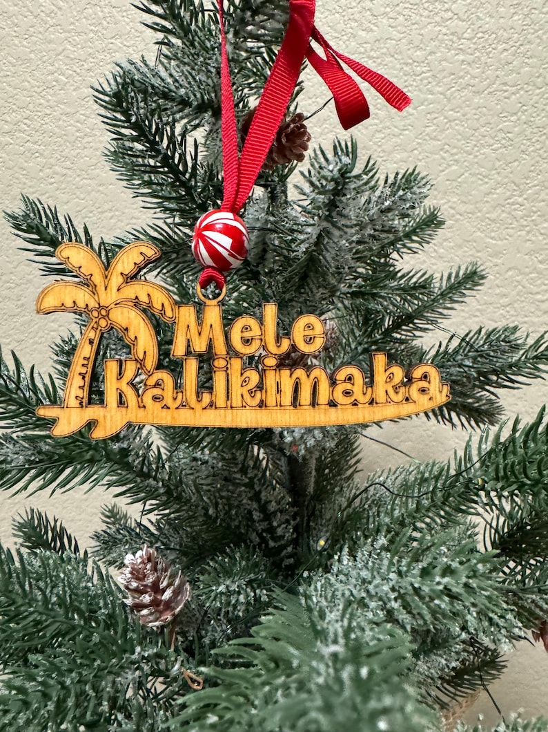 K&ouml;nnte beinhalten: Weihnachtsbaumschmuck aus Holz mit den Worten "Mele Kalikimaka" und einem Palmenmotiv. Der Schmuck h&auml;ngt an einem roten Band und ist mit einer rot-wei&szlig; gestreiften Kugel verziert. Der Schmuck wird auf einem Weihnachtsbaum pr&auml;sentiert.