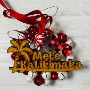 K&ouml;nnte beinhalten: Ein h&ouml;lzerner Weihnachtsschmuck mit den Worten "Mele Kalikimaka" und einem Palmen-Design. Der Schmuck ist von wei&szlig;en und roten Glocken, rotem Band und einer rot-wei&szlig; gestreiften Gl&uuml;hbirne umgeben.