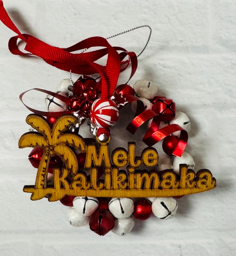 K&ouml;nnte beinhalten: Ein h&ouml;lzerner Weihnachtsschmuck mit den Worten "Mele Kalikimaka" und einem Palmen-Design. Der Schmuck ist von wei&szlig;en und roten Glocken, rotem Band und einer rot-wei&szlig; gestreiften Gl&uuml;hbirne umgeben.
