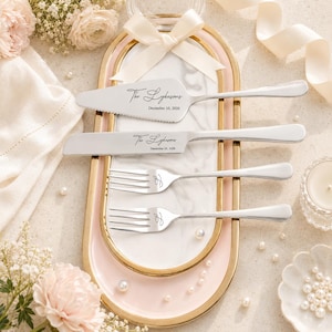 May include: A silver cake serving set consisting of a serrated cake knife and a cake server, both engraved with "The Lykasons" and "December 10, 2026", alongside two matching forks engraved with the letter "L", all resting on an oval marble tray.