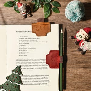 Puede incluir: Una p&aacute;gina de libro de cocina con un l&aacute;piz, sujeta por marcadores de cuero marr&oacute;n y rojo. Los marcadores presentan un dise&ntilde;o de reno y Pap&aacute; Noel. Los adornos navide&ntilde;os est&aacute;n dispuestos alrededor de la p&aacute;gina.