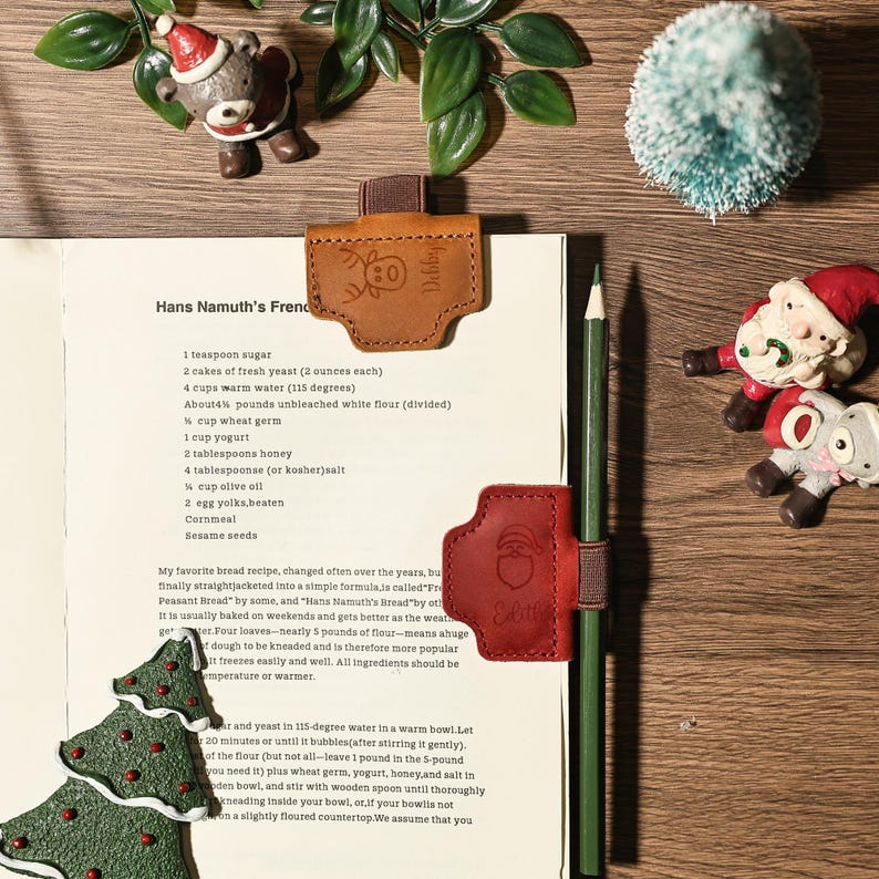 Puede incluir: Una p&aacute;gina de libro de cocina con un l&aacute;piz, sujeta por marcadores de cuero marr&oacute;n y rojo. Los marcadores presentan un dise&ntilde;o de reno y Pap&aacute; Noel. Los adornos navide&ntilde;os est&aacute;n dispuestos alrededor de la p&aacute;gina.