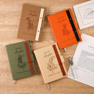 Puede incluir: Cuatro diarios de cuero en varios colores: marrón, naranja, verde y crema. Cada diario presenta el texto "THE NEXT chapter" y un diseño floral con un nombre debajo. Los diarios tienen un cierre de correa y una cinta de marcador.