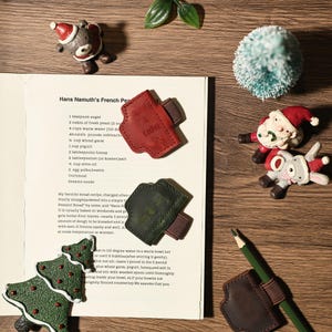 Puede incluir: Una colecci&oacute;n de adornos navide&ntilde;os y portal&aacute;pices de cuero se colocan sobre una superficie de madera. Los adornos incluyen una figura de Pap&aacute; Noel, un oso y un &aacute;rbol de Navidad. Los portal&aacute;pices son rojos, verdes y marrones.