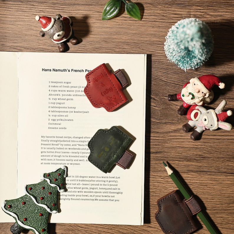 Puede incluir: Una colecci&oacute;n de adornos navide&ntilde;os y portal&aacute;pices de cuero se colocan sobre una superficie de madera. Los adornos incluyen una figura de Pap&aacute; Noel, un oso y un &aacute;rbol de Navidad. Los portal&aacute;pices son rojos, verdes y marrones.