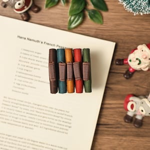 Puede incluir: Una colecci&oacute;n de seis sujetap&aacute;ginas de cuero de colores con bandas el&aacute;sticas marrones, dispuestos sobre una p&aacute;gina de texto. Los sujetap&aacute;ginas son en tonos marr&oacute;n, turquesa, naranja, rojo y verde. La p&aacute;gina est&aacute; sobre una superficie de madera, con adornos navide&ntilde;os.
