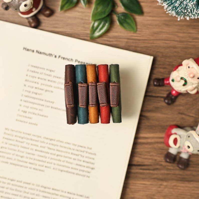 Puede incluir: Una colecci&oacute;n de seis sujetap&aacute;ginas de cuero de colores con bandas el&aacute;sticas marrones, dispuestos sobre una p&aacute;gina de texto. Los sujetap&aacute;ginas son en tonos marr&oacute;n, turquesa, naranja, rojo y verde. La p&aacute;gina est&aacute; sobre una superficie de madera, con adornos navide&ntilde;os.