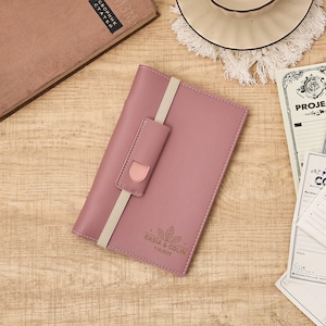 May include: A dusty rose-colored notebook with a cream-colored elastic band and a matching closure. The notebook has the names "BASIA & COLIN" and the date "9.10.2026" printed on the front. The notebook is lying on a wooden surface.