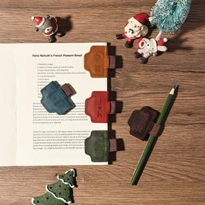 Puede incluir: Una vista superior de un libro de cocina abierto en una receta, con varios sujetap&aacute;ginas de cuero de varios colores. Cada soporte tiene una correa e iniciales. Tambi&eacute;n se ve un l&aacute;piz verde y adornos navide&ntilde;os.