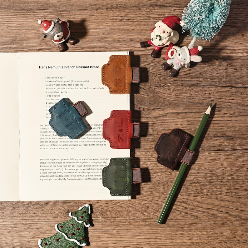 Puede incluir: Una vista superior de un libro de cocina abierto en una receta, con varios sujetap&aacute;ginas de cuero de varios colores. Cada soporte tiene una correa e iniciales. Tambi&eacute;n se ve un l&aacute;piz verde y adornos navide&ntilde;os.