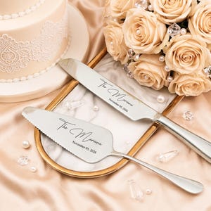 May include: A silver cake knife and server set engraved with "The Mariana" and the date November 05, 2026. The set rests on a gold-edged tray with a marble pattern, next to a bouquet of cream-coloured roses.