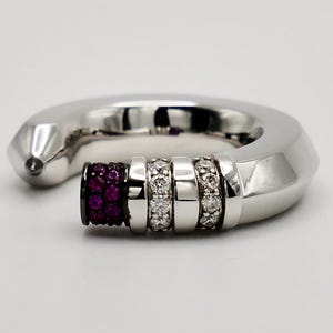 Pode incluir: Um anel prateado com um design geométrico. O anel apresenta uma secção de pequenas pedras redondas de cor vermelho escuro e três faixas de pedras claras e brilhantes. O anel tem um estilo moderno e elegante.