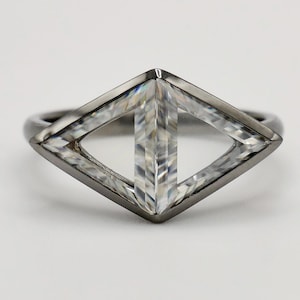 Puede incluir: Un anillo moderno con un diseño geométrico. El anillo presenta una banda gris oscuro y una pieza central transparente en forma de diamante con una barra vertical. La pieza central está hecha de múltiples facetas que captan la luz.