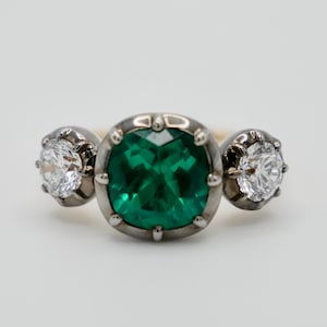 Puede incluir: Un anillo de tres piedras con una piedra central grande de color verde esmeralda y dos diamantes redondos brillantes más pequeños a cada lado.