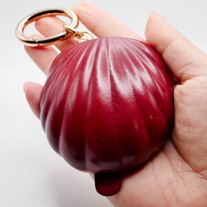 Op de afbeelding: Een dieprode, schelpvormige sleutelhanger met een goudkleurige ring. De sleutelhanger heeft een textuur met verticale ribbels. De sleutelhanger wordt in een hand gehouden tegen een witte achtergrond.