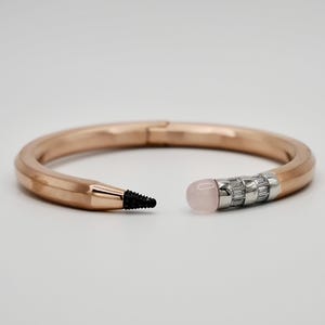 Op de afbeelding: Een roségouden armband ontworpen als een potlood. De armband heeft een zwarte grafietpunt aan één kant en een roze edelsteen gum met zilveren accenten aan de andere kant. De armband is open en toont het ontwerp. Omtrek ca. 18 cm.