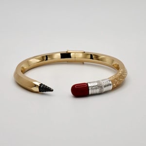 May include: A gold-coloured bangle bracelet designed to look like a pencil. The bracelet has a black tip at one end and a red eraser with silver accents at the other. The pencil-themed jewellery is a unique accessory, measuring approximately 18 cm in circumference.