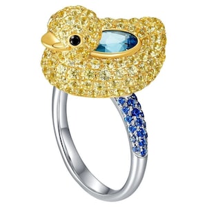 May include: A gold ring with a yellow duck design set with blue and yellow gemstones. The duck's head is turned to the side and its beak is open. The ring is set on a white background.