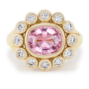 May include: A gold ring featuring a large, oval-cut pink gemstone surrounded by a halo of smaller, round, white gemstones. The ring has a classic, elegant design, perfect for special occasions.